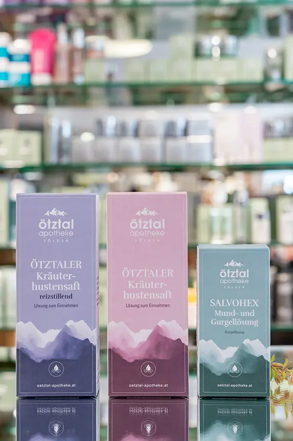 Auf einer Theke mit unscharfen Produktregalen im Hintergrund stehen drei Hautpflegeboxen mit minimalistischen Bergmotiven in Lila-, Rosa- und Grüntönen. Neben den Boxen steht eine kleine Pflanze. Es handelt sich um Produkte der Eigenherstellung.
