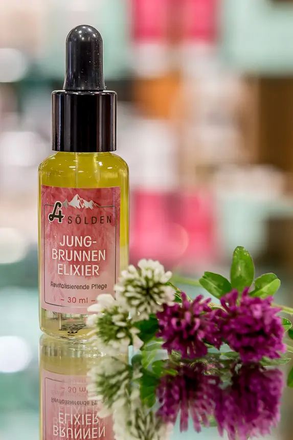 Ein kleines Glasfläschchen mit Gesichtsöl und einem Tropfverschluss steht neben einem Strauß frischer Blumen auf einer spiegelnden Oberfläche mit einem unscharfen bunten Hintergrund.