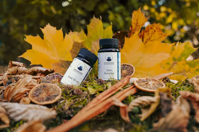 Zwei kleine weiße Flaschen mit schwarzen Verschlüssen stehen auf einem moosbewachsenen Boden, umgeben von getrockneten Blättern, Orangenscheiben, Zimtstangen und großen gelben Herbstblättern im Hintergrund.