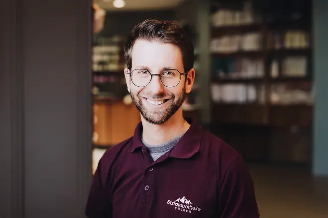 Ein lächelnder Mann mit Brille und Bart, der ein Polohemd trägt, steht in der Ötztal Apotheke, in der im Hintergrund Regale zu sehen sind.