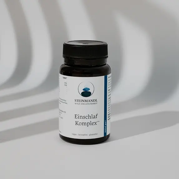 Eine schwarze Flasche mit weißem Etikett steht auf einer weißen Fläche, hinter der die Schatten eines Geländers oder von Stangen diagonale Linien erzeugen. Das Etikett enthält minimalistische Grafiken und Text.