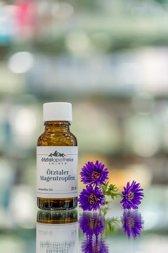 Eine kleine Braunglasflasche mit weißem Etikett steht neben zwei violetten Blumen auf einer glänzenden Oberfläche mit unscharfem Hintergrund.