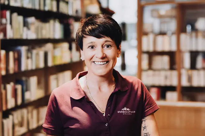Eine Person mit kurzen dunklen Haaren und einem Polohemd lächelt in der Apotheke, mit Regalen voller Arzneimittel im Hintergrund.