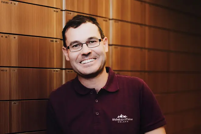 Ein lächelnder Mann mit Brille und einem Poloshirt steht vor einem Holzschrank.