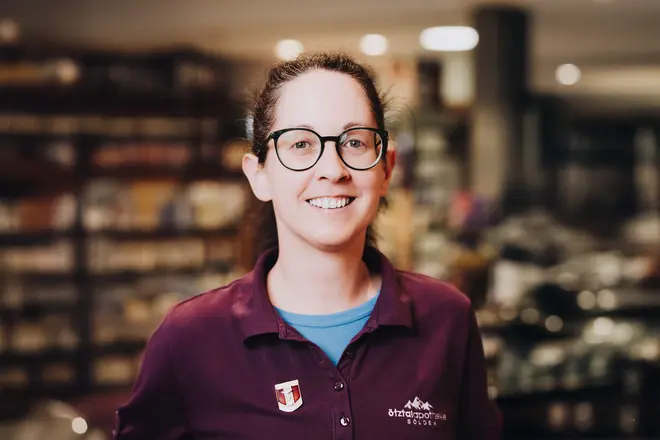 Eine Frau mit Brille und zurückgebundenem dunklem Haar, die ein Oberteil trägt, steht lächelnd in der Apotheke mit Regalen im Hintergrund.