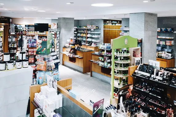Eine hell erleuchtete Apotheke mit Regalen, die mit verschiedenen Produkten bestückt sind, darunter Arzneimittel, Kosmetikartikel und Körperpflegeartikel.